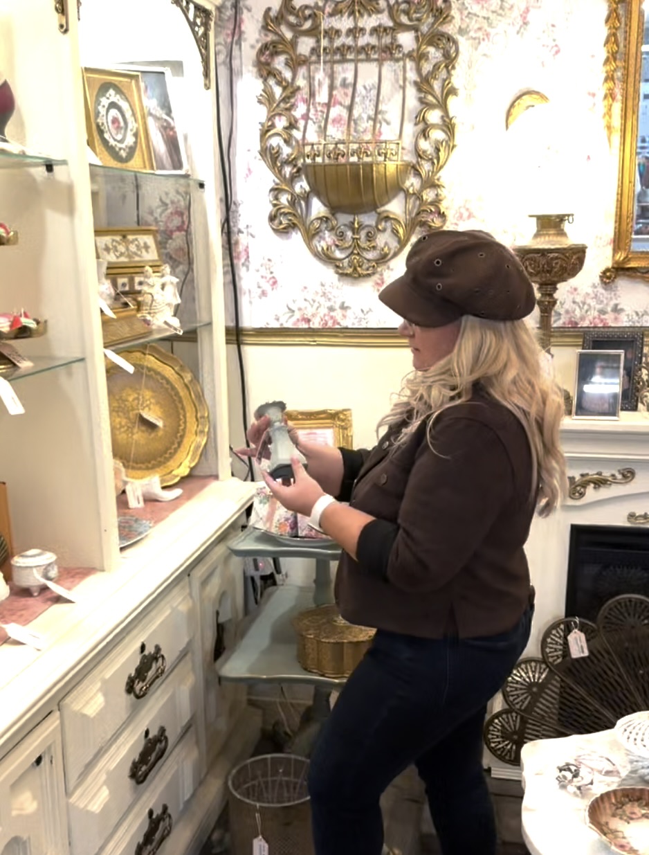 Marissa, in a brown cap, looks closely at a vintage candlestick in an antique shop with gold-framed mirrors and floral wallpaper